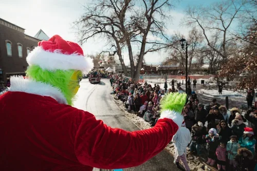 A person dressed as the Grinch in a Santa outfit waving to a line of people in winter clothing along a snow-covered street during a holiday parade.