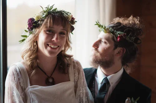 A smiling woman and a bearded man with curly hair, both wearing flower crowns, in a warmly lit room.