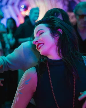 A young woman dancing and smiling in a crowded, colorful party or club, illuminated by neon lights.