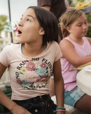 Two young girls sitting at a table outdoors, one girl looking surprised or excited, wearing a beige t-shirt with floral design, the other girl wearing a pink dress and holding a plate.