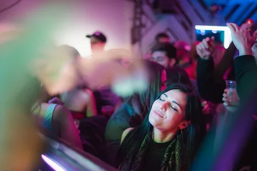 A young woman with dark hair and closed eyes enjoying herself at a crowded party or concert with colorful lighting.