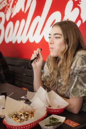 Young woman eating food at a restaurant, sitting at a table with basket of nachos and jalapenos, with a red wall and large white text in the background.