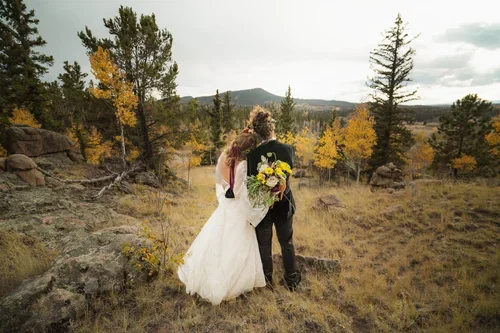 Bride and groom standing outdoors in a fall landscape, with trees and hills in the background, the bride holding a bouquet of flowers.
