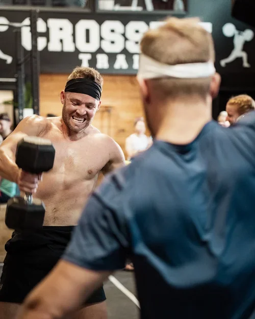 Two men wearing headbands are competing in a fitness challenge, with one holding a dumbbell and flexing his bicep at CrossFit gym.
