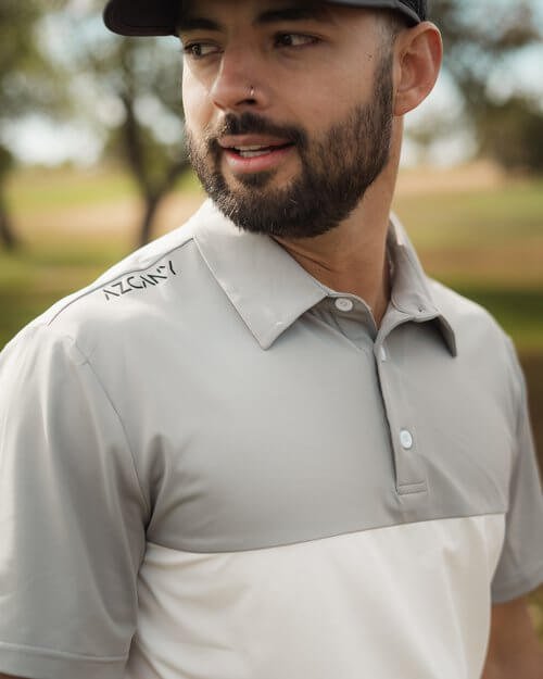 A man with a beard  wearing a light gray collared golf shirt outdoors.