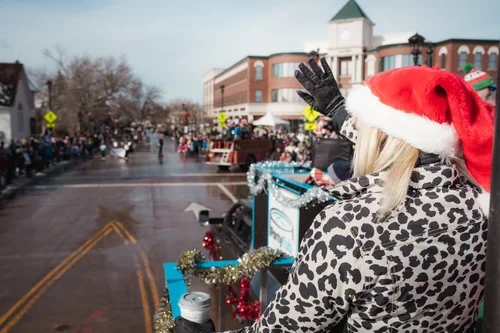 A person wearing a Santa hat and leopard print coat waving from a decorated parade float during a Christmas parade in a small town.