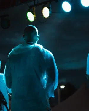 A person with a shaved head wearing a light-colored shirt standing on a stage with colorful stage lights overhead.