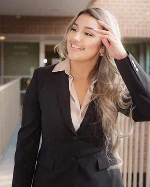 A young woman with long, wavy blonde hair smiling and touching her forehead, dressed in a black blazer and white blouse, standing outdoors in front of a brick building.