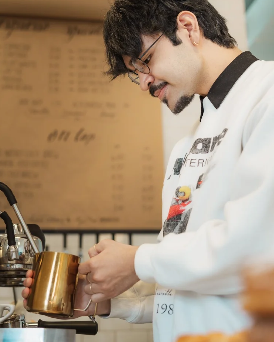 A man with glasses, dark hair, and a beard, smiling, standing in a coffee shop, holding a gold-colored mug, preparing a drink.