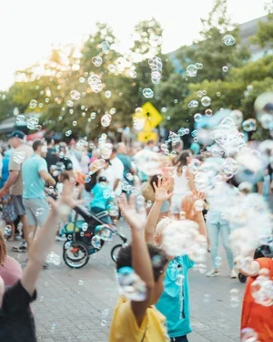 Children and families playing with bubbles outdoors in a park, with trees and a wheelchair in the background