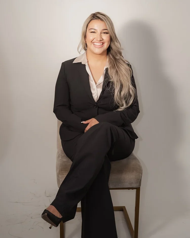 A woman in a black suit and white blouse sitting on a beige chair, smiling and posing for the camera.