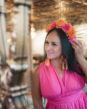 Woman wearing a pink dress and a floral crown with pink and orange roses, holding her hair back with her right hand, outdoors with blurred background.