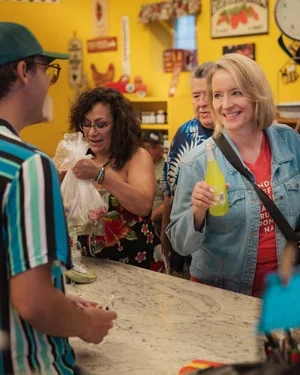Four people at a colorful store counter, woman holding a yellow bottle, smiling, others browsing and smiling.