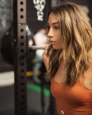 A young woman with long, wavy hair in a gym or fitness setting, standing near a black metal rack.