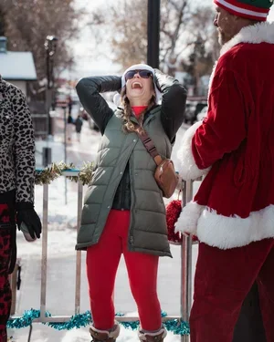 A woman in winter clothing laughing with her hands on her head, standing outdoors in a snowy setting, with a person dressed as Santa Claus in the foreground.