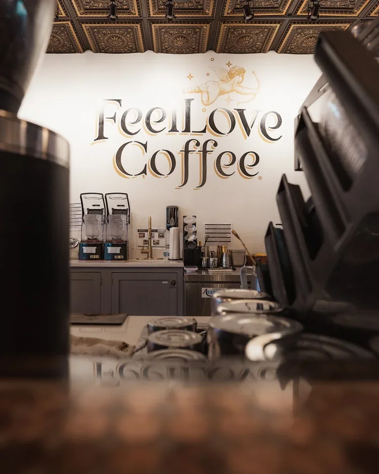 Interior of a coffee shop with a wall sign that reads 'Feel Love Coffee' and a decorative angel illustration above it, with coffee machines and supplies visible in the background.