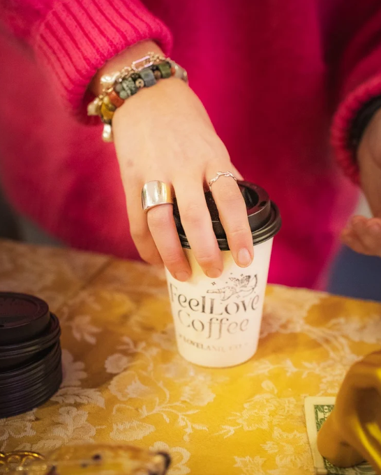 A person wearing a red sweater and multiple bracelets, holding a disposable coffee cup labeled "Feel Love Coffee" with a black lid, on a yellow floral tablecloth.