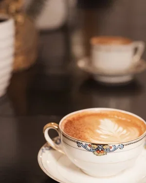 A cup of coffee with foam art on a saucer, sitting on a dark surface with a blurred background.