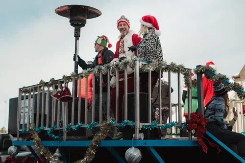 People on a festive parade float decorated with Christmas garlands, wearing holiday costumes including Santa hats.