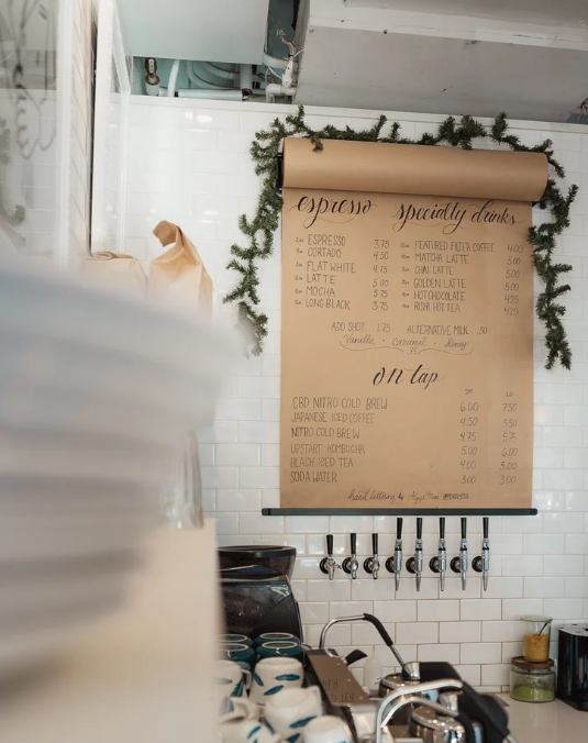 A menu board in a coffee shop listing espresso drinks and on-tap beverages, decorated with pine branches.