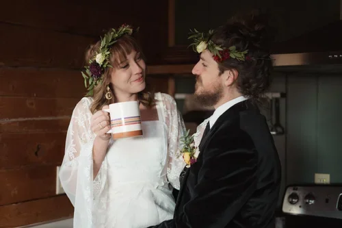 Couple dressed in wedding attire with flower crowns, woman holding a mug, in a cozy indoor setting.