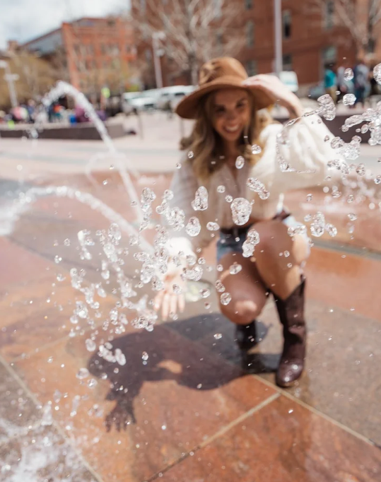 Woman wearing a hat and boots kneeling near a splash of water in an outdoor urban setting.