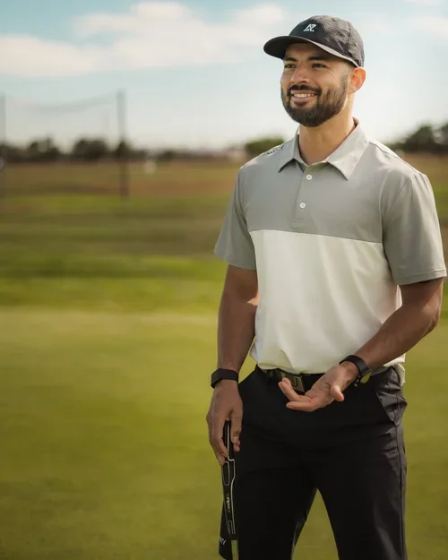 A man with a beard and a cap standing on a golf course under a partly cloudy sky, holding a golf club.