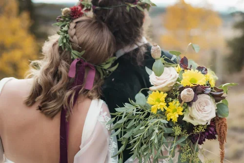 A couple at a wedding, with the woman wearing a floral crown and the man holding a bouquet of yellow and purple flowers, outdoors during fall.