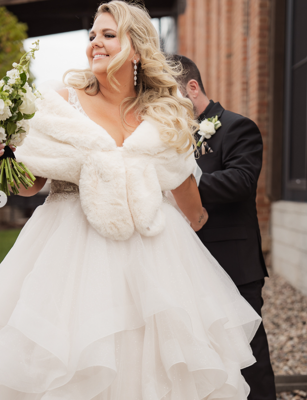 A bride with long blonde hair in a white wedding gown and fur shawl, smiling and holding a bouquet, standing outside a building. A groom in a black suit with a white boutonniere stands in the background.