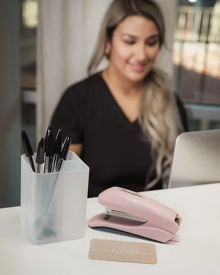A woman sitting at a desk with a white container of black pens, a pink stapler, a closed laptop, and a business card.