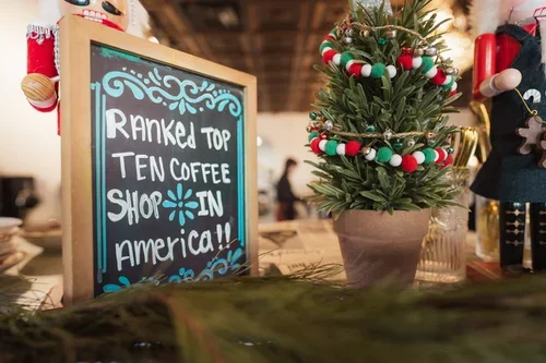 A Christmas tree decorated with red, white, and green ornaments next to a chalkboard sign that reads 'Ranked top ten coffee shop in America,' inside a cozy cafe.
