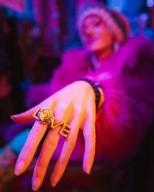 A person wearing a purple fur coat showing their hand with two gold rings, one with a skull and one with a rectangle, against a colorful background.