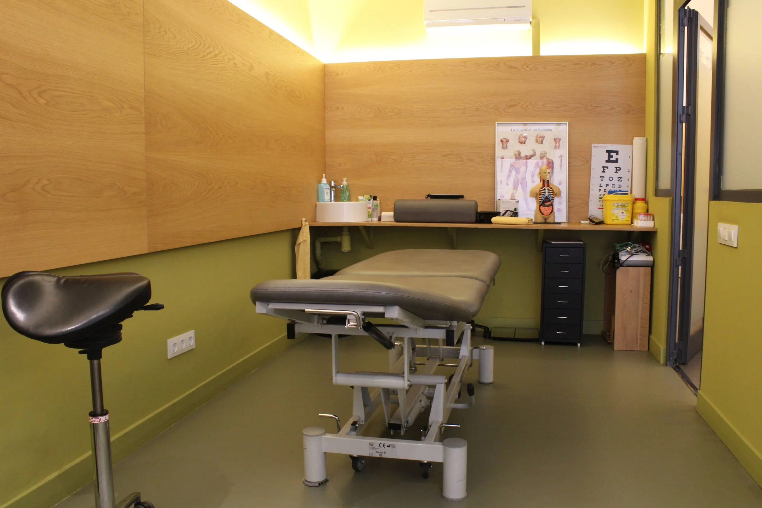 Medical examination room with an adjustable examination table, a stool, and medical supplies on a counter, including anatomical posters and an eye chart.