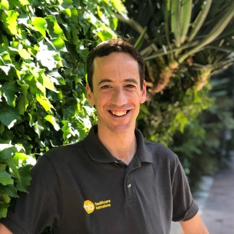 A smiling man wearing a black polo shirt with a logo, standing outdoors in front of lush green foliage.