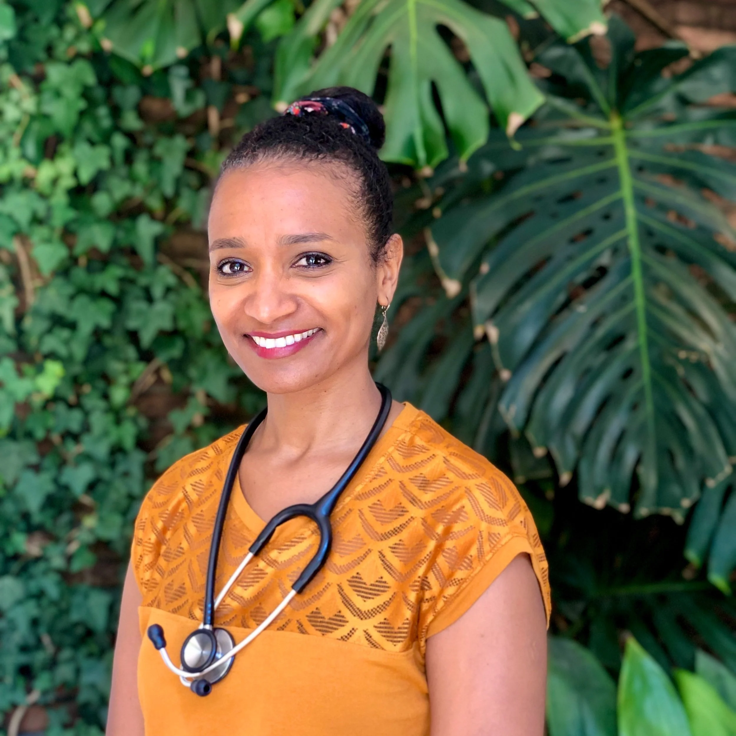 A smiling woman with dark hair in a bun, wearing a yellow top and a stethoscope around her neck, standing outdoors with broad green tropical leaves in the background.