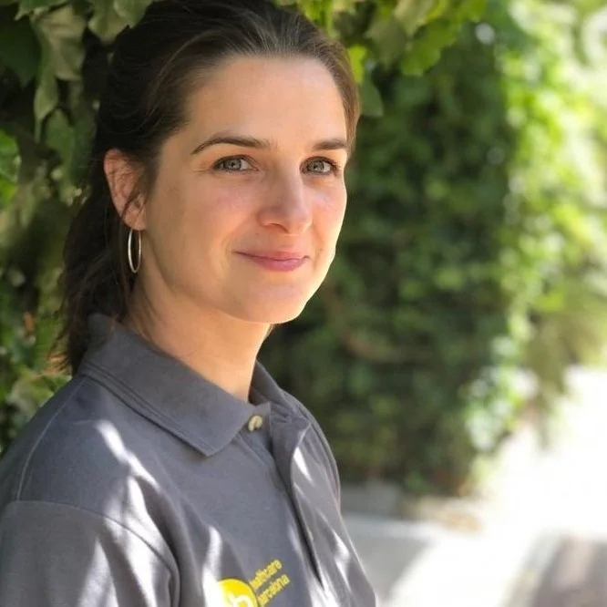 A woman with dark brown hair tied back, wearing hoop earrings and a gray polo shirt, standing outdoors next to green foliage, smiling gently at the camera.