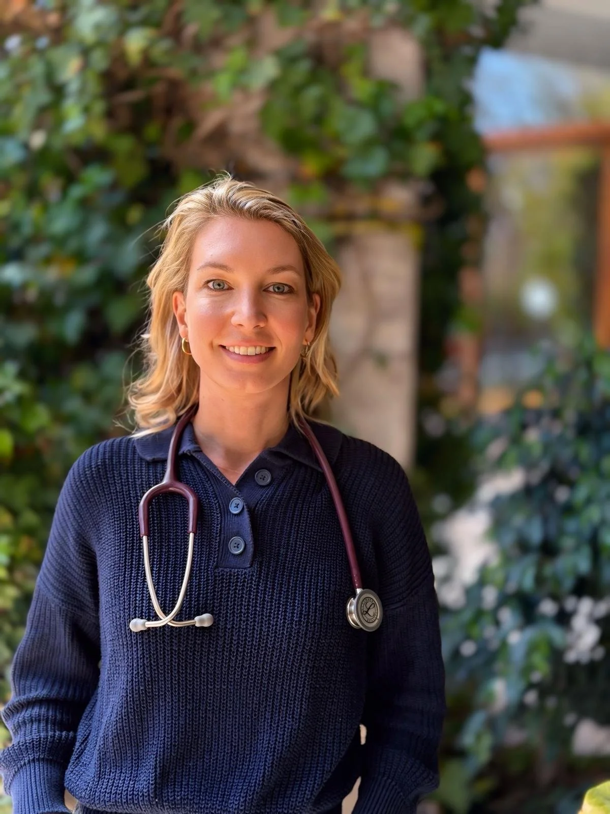 A smiling female healthcare professional with blonde hair, wearing a navy blue sweater and a stethoscope around her neck, standing outdoors with green foliage in the background.