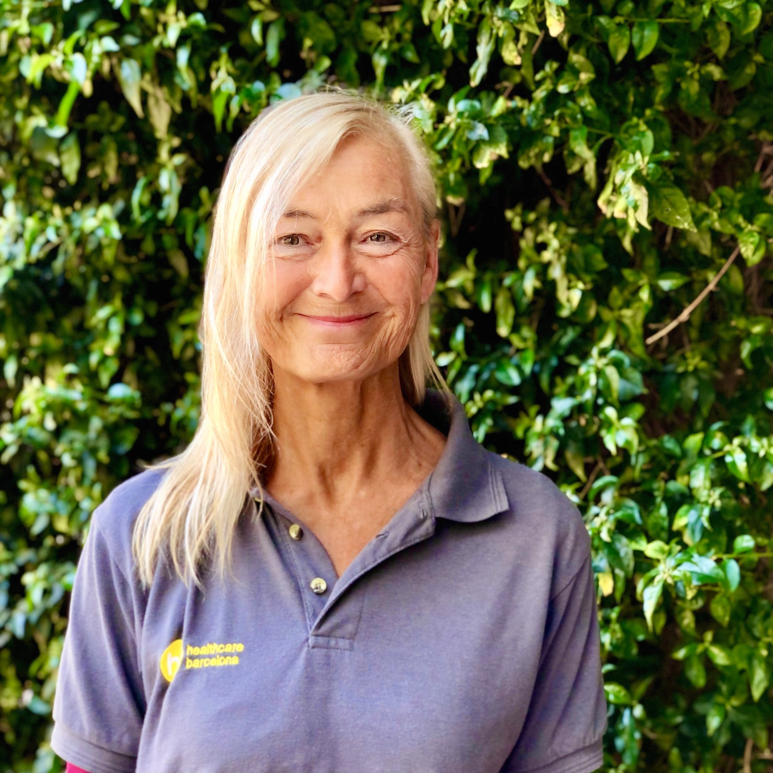 Smiling older woman with long blonde hair wearing a gray polo shirt with yellow text, standing in front of a green leafy background.