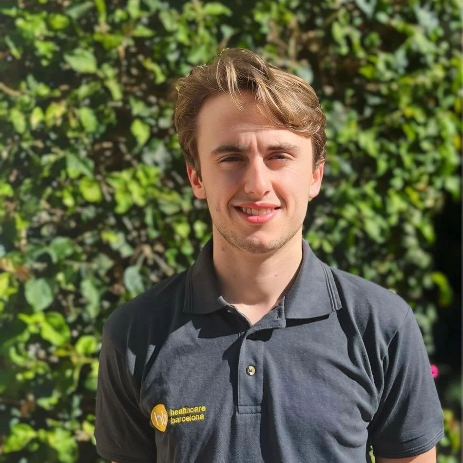 A young man with light brown hair and fair skin, wearing a dark gray polo shirt with a healthcare barcelona logo, standing outdoors in front of green foliage.