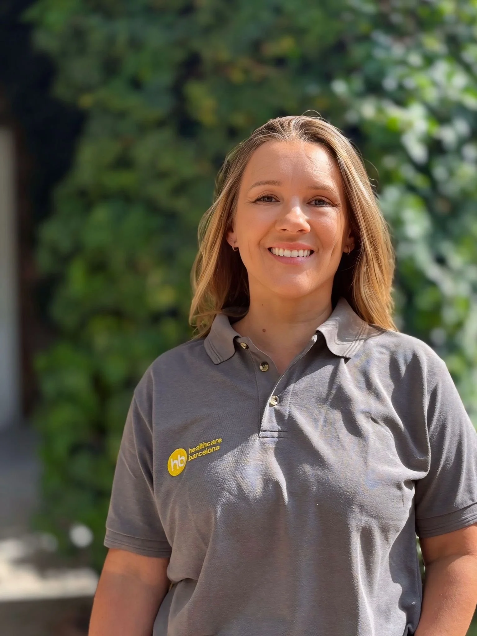A woman wearing a gray polo shirt with a 'healthcare barcelona' logo, smiling outdoors with green trees in the background.