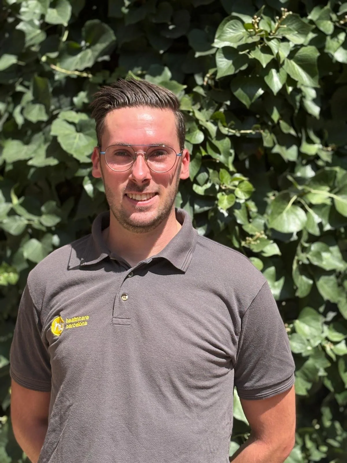 A man wearing glasses and a gray polo shirt with yellow logo stands outdoors in front of leafy green bushes.