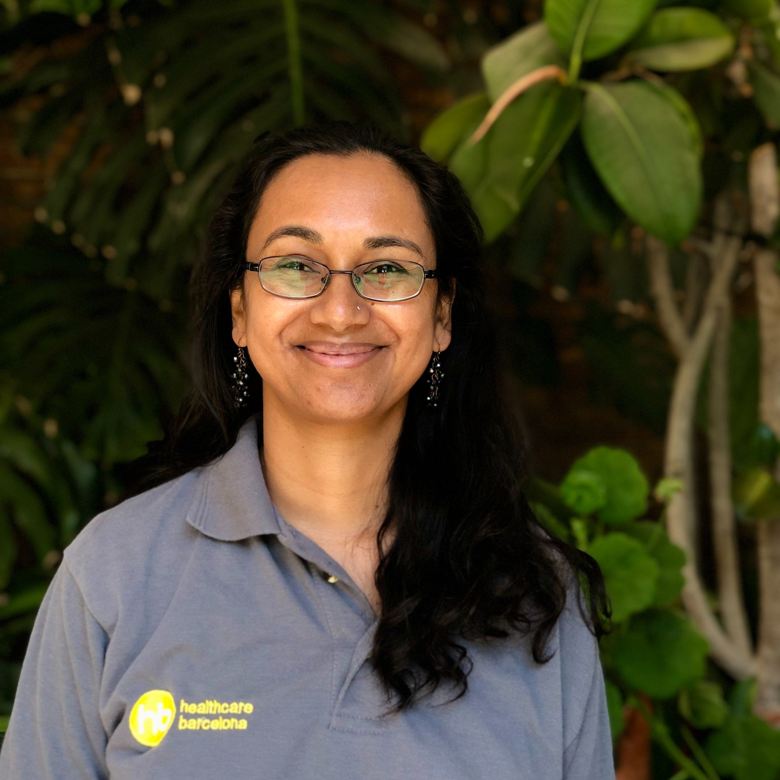 A woman with long dark hair, glasses, and earrings, smiling and wearing a gray polo shirt with a yellow and white 'healthcare barcelona' logo, standing outdoors with green foliage in the background.