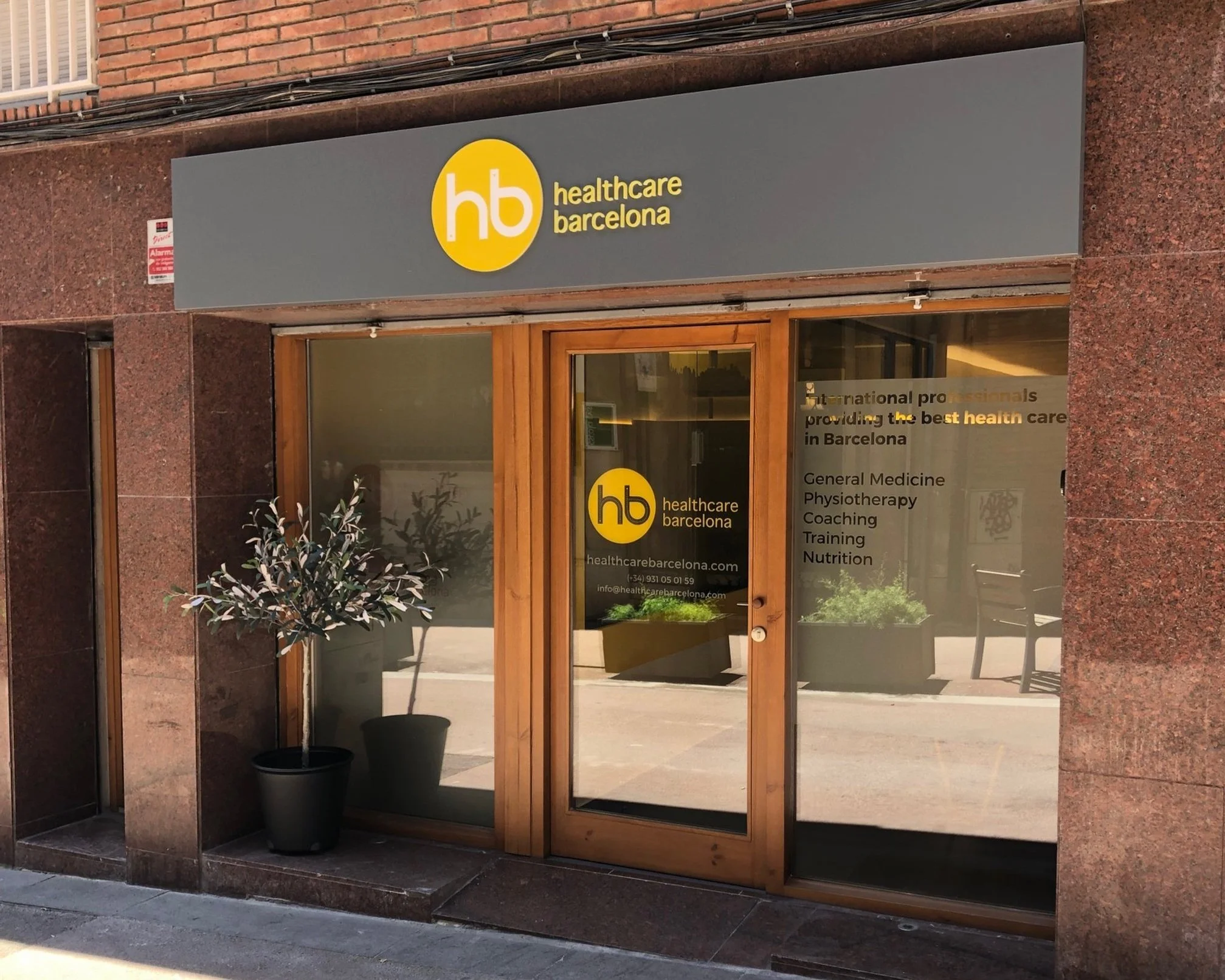 Exterior of a healthcare clinic with wooden door and glass window, displaying the logo 'hb healthcare barcelona' on the door and sign, and a potted plant outside.