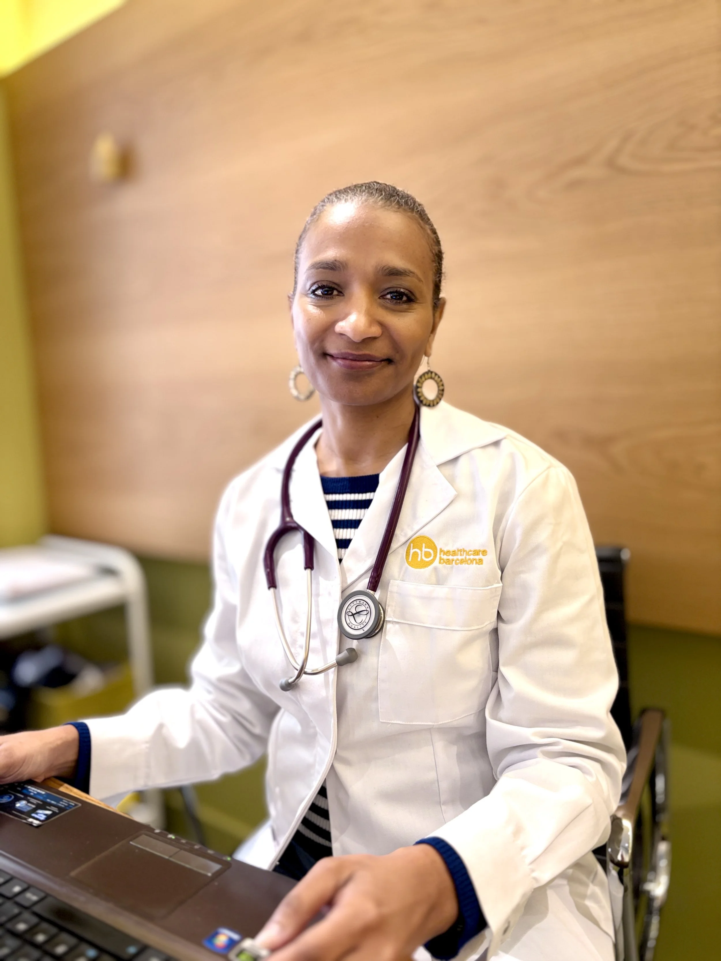 Healthcare professional in white coat with stethoscope, sitting at desk, smiling at camera, in medical office.