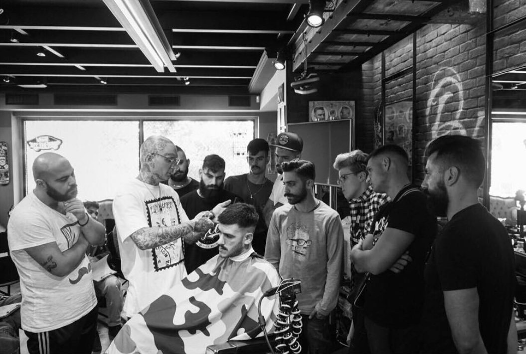 Barber giving haircut to a man while a group of men watch in a modern barbershop.