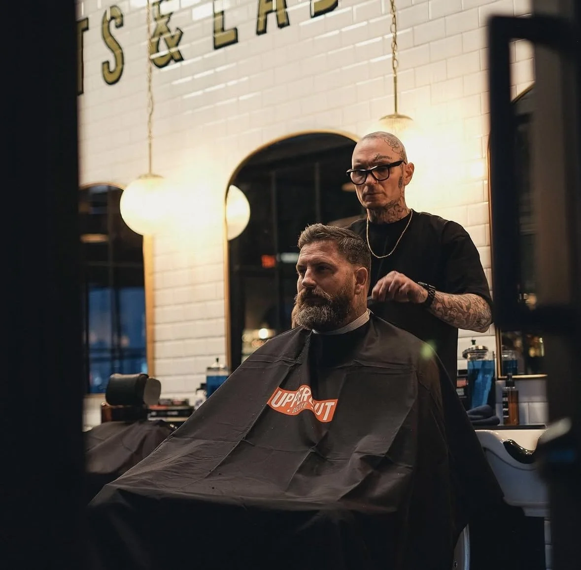 A man getting a haircut at a barbershop, sitting in a chair with a black cape, while a barber with tattoos and glasses styles his hair. The barbershop has white brick walls and warm lights.