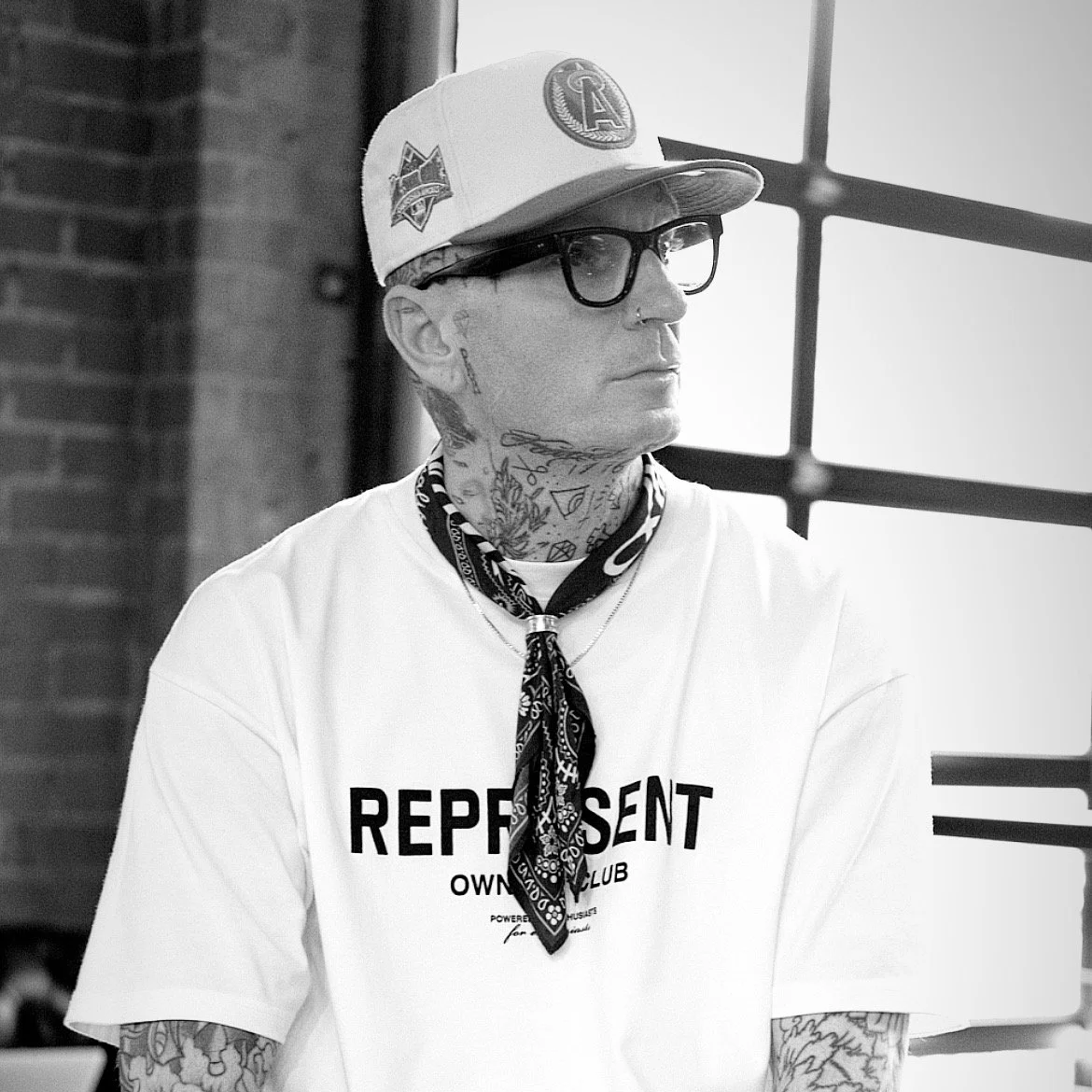 A tattooed man wearing glasses, a cap, and a bandana around his neck, sitting indoors near a large window with brick wall in the background.