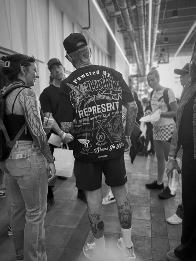 A man with tattoos on his arms and legs, wearing a cap, sunglasses, a graphic T-shirt, shorts, and sneakers, standing with a group of women in an indoor public space.
