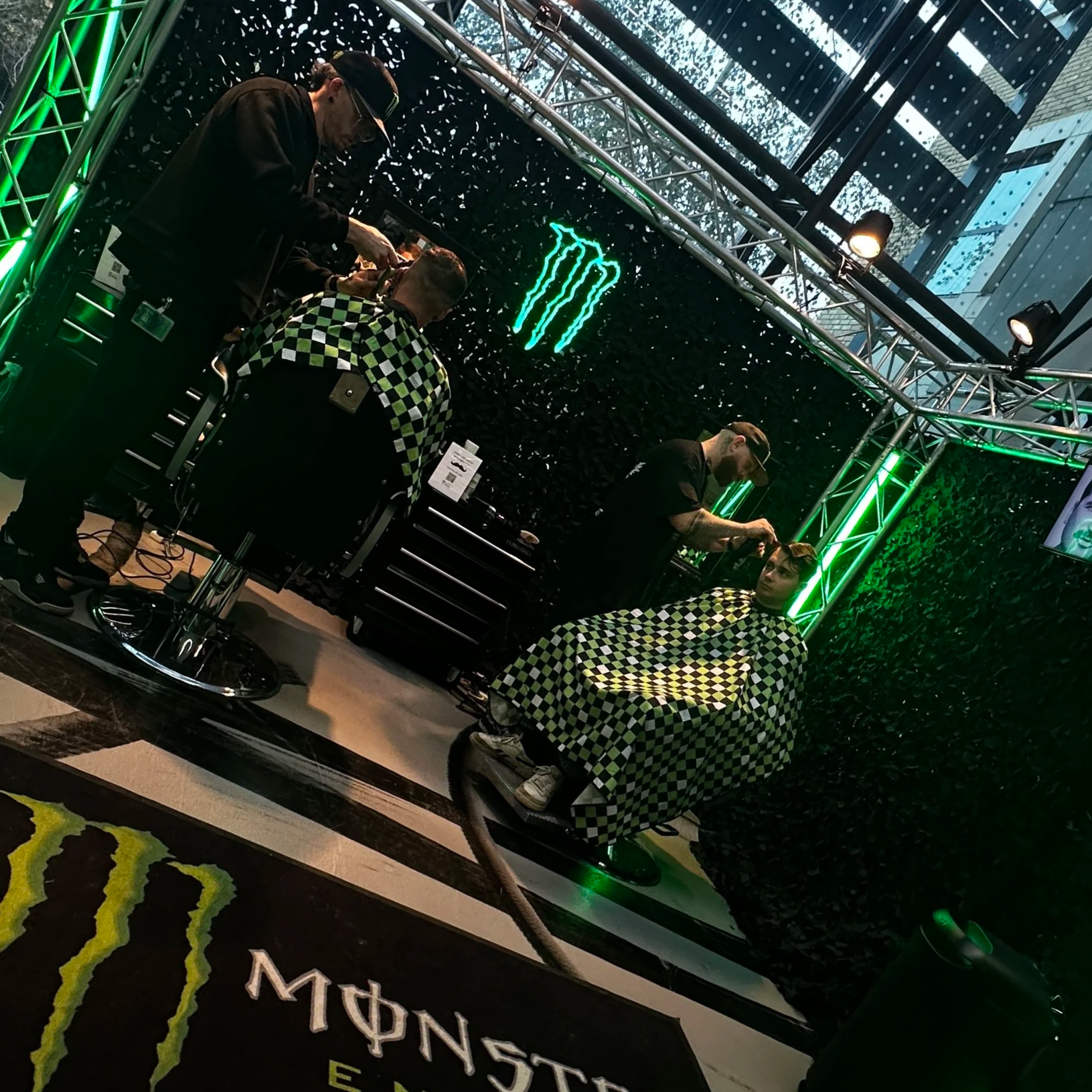 A barbershop setup inside a store featuring a black wall with green neon Monster logo, lime green neon lights framing the area, and a person getting a haircut with a barber doing the haircut. The customer is seated in a barber chair covered with a black and lime green checkered cape.