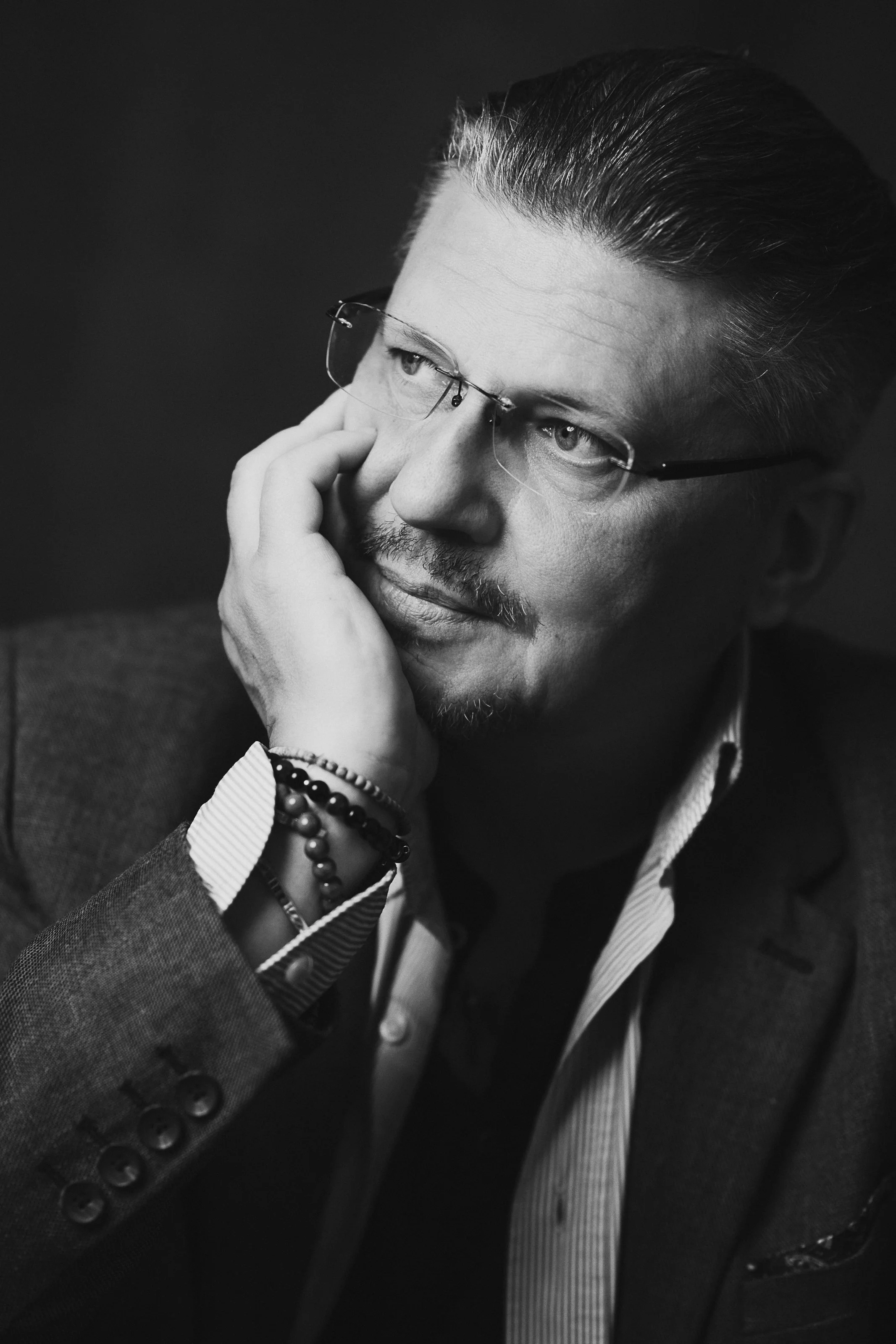 A black and white photo of a man with slicked-back hair, glasses, a mustache, and a goatee. He is wearing a suit jacket, a striped shirt, and a beaded bracelet, and is resting his face on his hand.